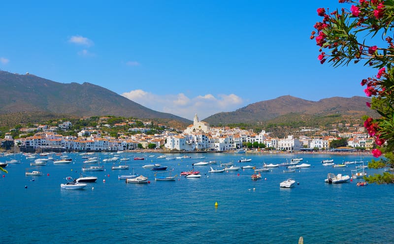 Casas brancas de Cadaqués descendo em socalcos até ao porto, barcos ancorados na baía calma e as rochas do Cap de Creus ao fundo, sob luz de fim de tarde.