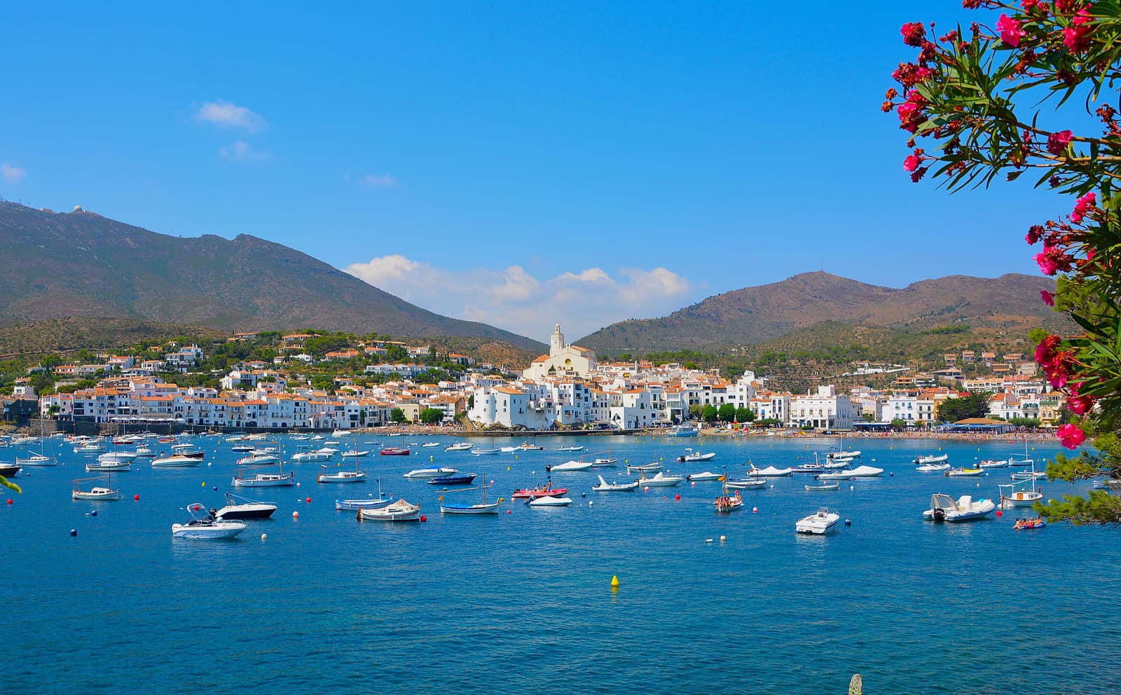 Casas brancas de Cadaqués descendo em socalcos até ao porto, barcos ancorados na baía calma e as rochas do Cap de Creus ao fundo, sob luz de fim de tarde.
