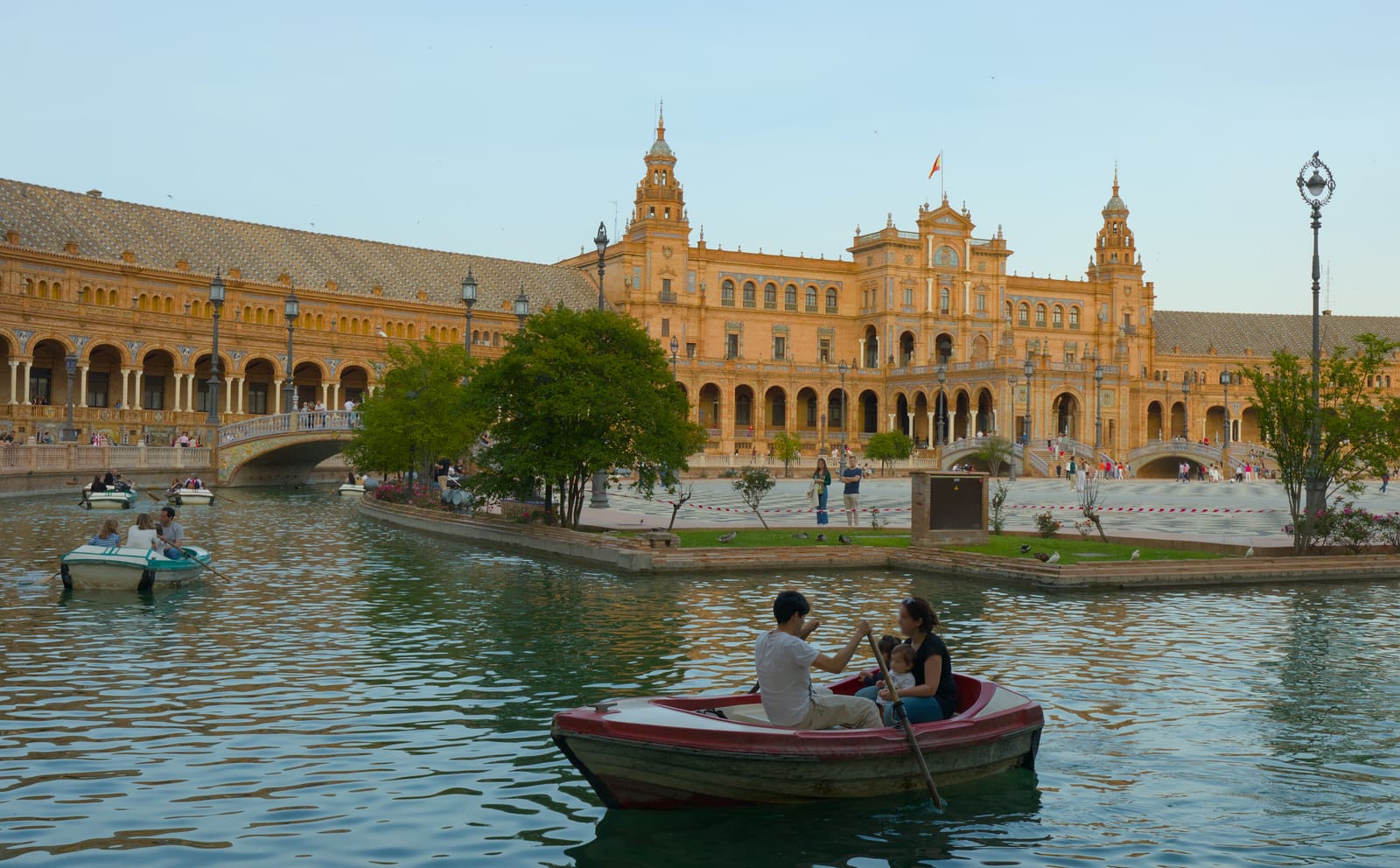 Casal remando num barco no canal da Plaza de España, em Sevilha, com o edifício de tijolo e as torres ao fundo